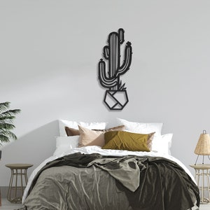 May include: Black metal wall art featuring a cactus in a geometric pot. The art is mounted on a white wall above a bed with a green blanket and pillows.