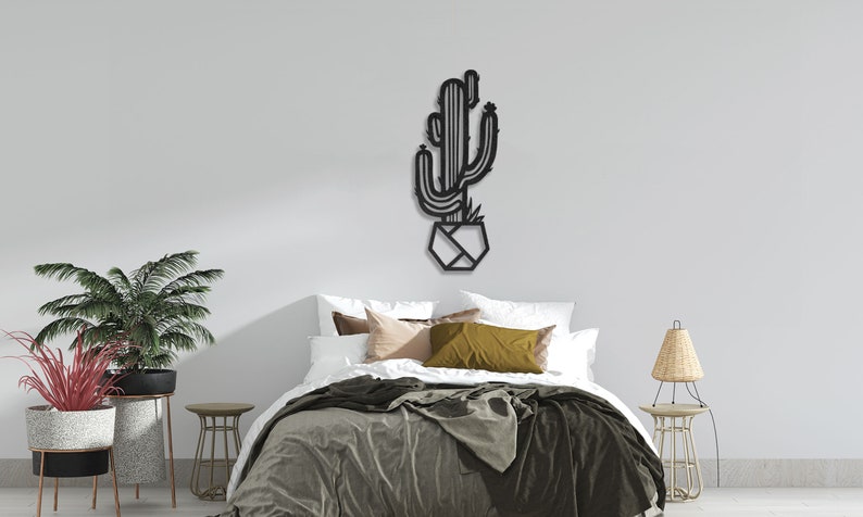 May include: Black metal wall art featuring a cactus in a geometric pot. The art is mounted on a white wall above a bed with a green blanket and pillows.