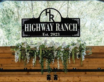 May include: A black metal sign hanging from chains, featuring the letters HR in an arch, the text HIGHWAY RANCH, and Est. 2023. The sign has decorative scrollwork on the corners and is positioned above a wooden wall adorned with white flowers and greenery.