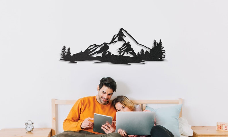 May include: A couple sits on a bed in front of a black metal wall decor depicting a mountain range with trees. The couple is looking at a tablet and a laptop.