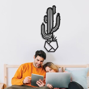 May include: A couple sit on a bed, looking at a tablet and laptop. A black metal cactus wall decor hangs above them.