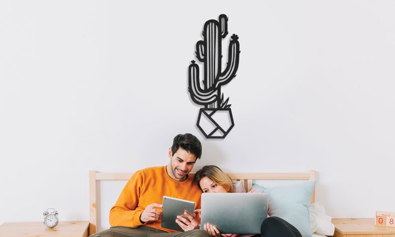 May include: A couple sit on a bed, looking at a tablet and laptop. A black metal cactus wall decor hangs above them.