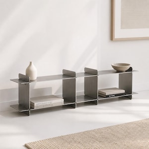 Op de afbeelding: Een modern, zilverkleurig metalen rek met een minimalistisch ontwerp. De unit heeft meerdere planken en verticale steunen. Het is gestyled met een witte vaas, een beige kom en twee boeken met de titels "Remote Places to Stay" en "Rivers".