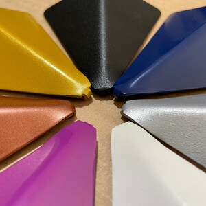 May include: A collection of eight triangular-shaped objects in various colors, including gold, black, navy blue, gray, white, purple, copper, and a textured gray. The objects are arranged in a circular pattern.