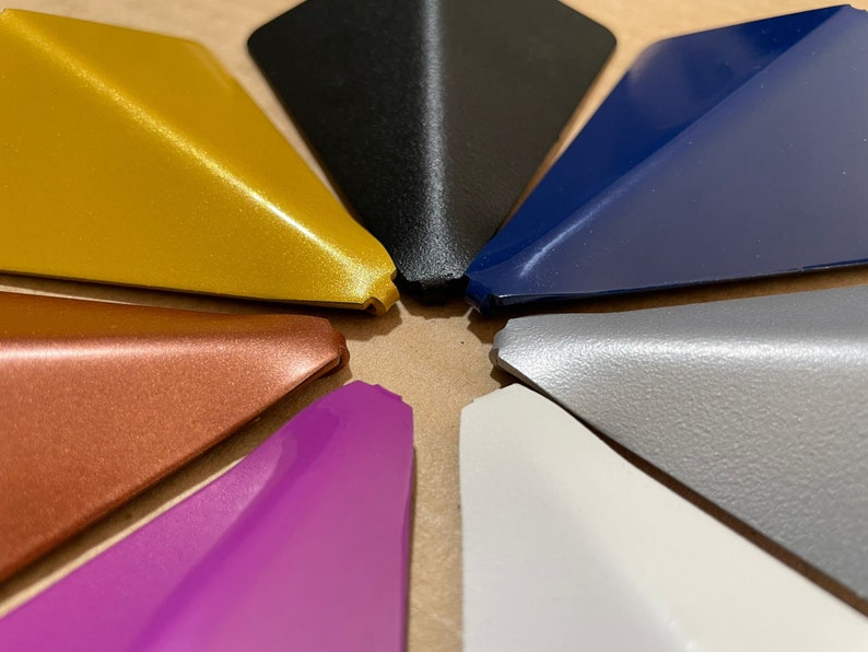 May include: A collection of eight triangular-shaped objects in various colors, including gold, black, navy blue, gray, white, purple, copper, and a textured gray. The objects are arranged in a circular pattern.