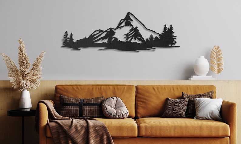 May include: Black metal wall art depicting a mountain range with snow-capped peaks and pine trees. The art is mounted on a white wall above a brown couch with pillows.
