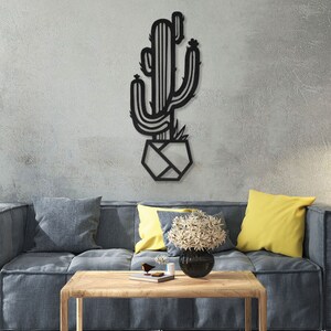 May include: Black metal wall art featuring a cactus in a geometric pot. The cactus is stylised with a simple design and has a few small flowers on top. The art is hanging on a grey wall in a living room setting.