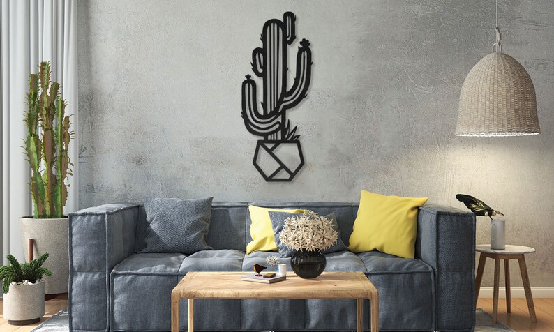 May include: Black metal wall art featuring a cactus in a geometric pot. The cactus is stylised with a simple design and has a few small flowers on top. The art is hanging on a grey wall in a living room setting.