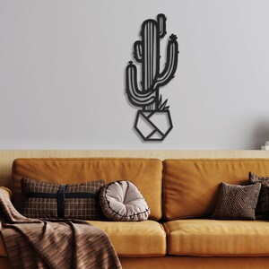 May include: Black metal wall art depicting a stylised cactus in a geometric pot. The cactus is minimalist in design, with a single stem and multiple arms. The art is mounted on a white wall above a brown sofa with cushions.