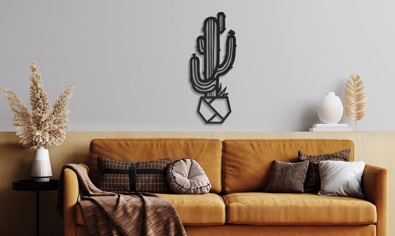 May include: Black metal wall art depicting a stylised cactus in a geometric pot. The cactus is minimalist in design, with a single stem and multiple arms. The art is mounted on a white wall above a brown sofa with cushions.