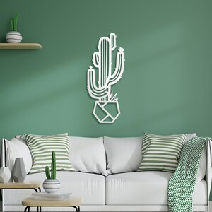 May include: White metal wall art depicting a cactus plant in a geometric pot. The cactus is outlined in a simple, modern style. The art is displayed on a green wall in a living room setting.