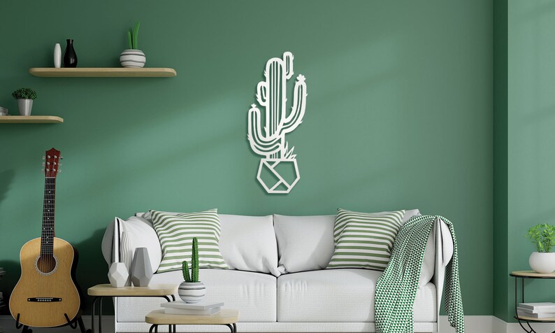 May include: White metal wall art depicting a cactus plant in a geometric pot. The cactus is outlined in a simple, modern style. The art is displayed on a green wall in a living room setting.