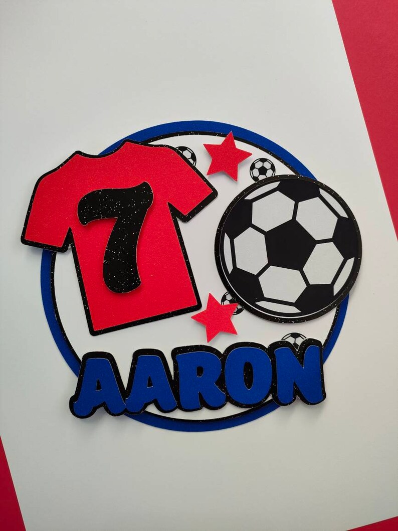 May include: A red soccer jersey with the number 7 on it, a black and white soccer ball, two red stars, and the name "AARON" in blue letters on a white background.