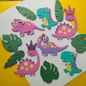May include: A collection of die-cut dinosaur shapes in pink, purple, and teal, featuring yellow spots and glittery accents like bows and crowns, accompanied by several green glittery tropical leaf cutouts arranged on a white surface.