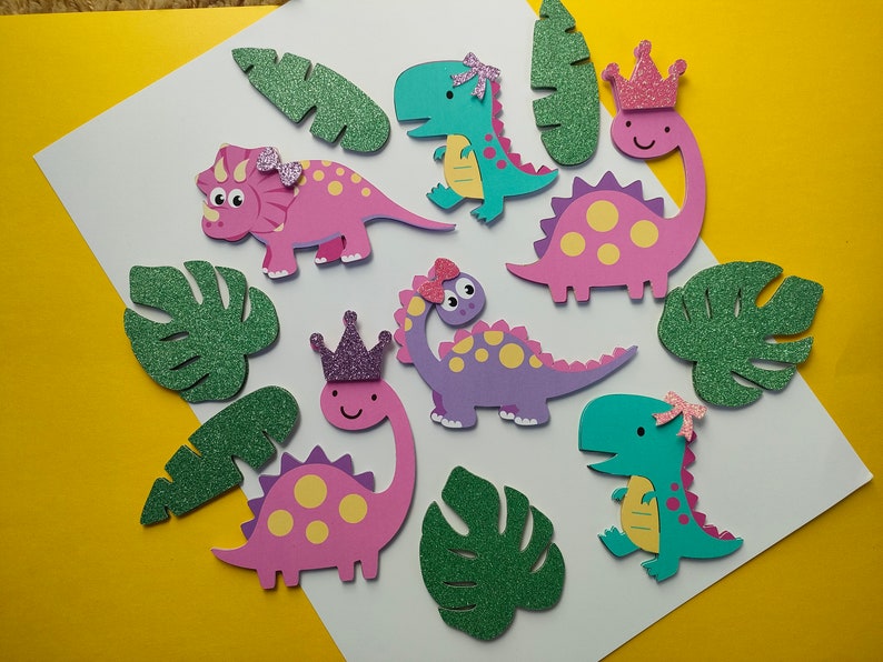 May include: A collection of die-cut dinosaur shapes in pink, purple, and teal, featuring yellow spots and glittery accents like bows and crowns, accompanied by several green glittery tropical leaf cutouts arranged on a white surface.