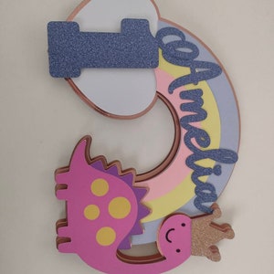 May include: A layered cake topper featuring the number one in purple glitter, a pastel rainbow, and a pink dinosaur wearing a gold glitter crown. The name Amelia is written in purple glitter script across the rainbow, with rose gold edges.