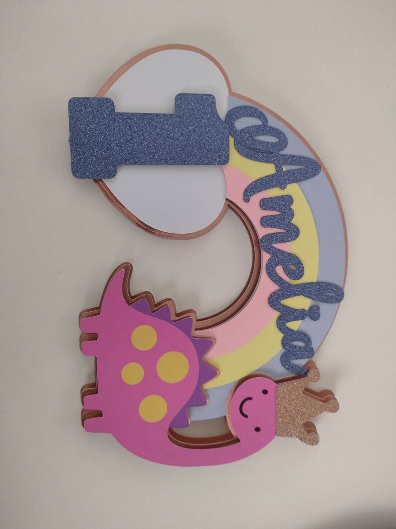 May include: A layered cake topper featuring the number one in purple glitter, a pastel rainbow, and a pink dinosaur wearing a gold glitter crown. The name Amelia is written in purple glitter script across the rainbow, with rose gold edges.