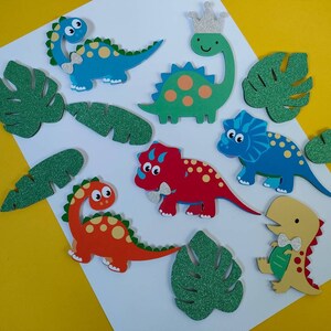 May include: A collection of flat, die-cut dinosaur and tropical leaf shapes. The dinosaurs are blue, green, red, and yellow, some with glittery silver crowns or bow ties. The leaves are green with a glittery texture, arranged on a white surface.