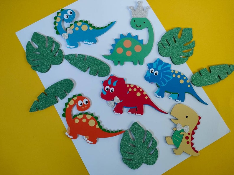 May include: A collection of flat, die-cut dinosaur and tropical leaf shapes. The dinosaurs are blue, green, red, and yellow, some with glittery silver crowns or bow ties. The leaves are green with a glittery texture, arranged on a white surface.