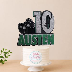 May include: A cake topper featuring a black gaming controller silhouette next to the number 10 in silver glitter, with the name AUSTEN in green glitter below. The topper is displayed on a white cake on a white stand with a GMs Crafty Creations logo.