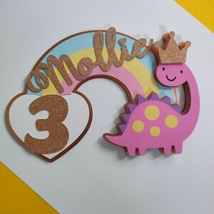 May include: A decorative cake topper featuring a pastel rainbow, a white heart with a glittery gold number 3, and a pink dinosaur wearing a glittery gold crown. The name Mollie is written in glittery gold script across the rainbow arch.