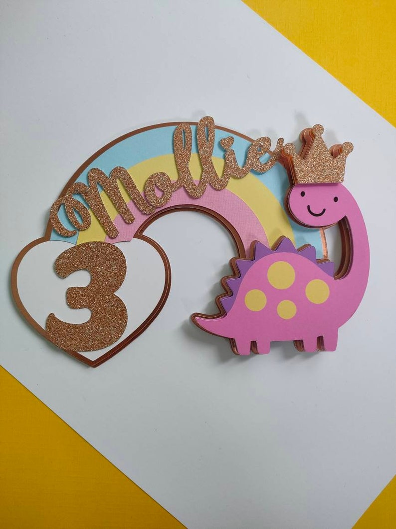 May include: A decorative cake topper featuring a pastel rainbow, a white heart with a glittery gold number 3, and a pink dinosaur wearing a glittery gold crown. The name Mollie is written in glittery gold script across the rainbow arch.