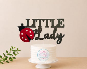 May include: A black glittery cake topper featuring the text Little Lady in a combination of block and script fonts. To the left of the text is a red ladybug with black spots and black antennae. The topper is mounted on a clear stick on a white cake.