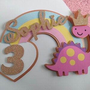 May include: A layered cake topper featuring a pastel rainbow, a white heart with a glittery gold number 3, and a pink dinosaur wearing a glittery gold crown. The name Sophie is written in glittery gold script across the rainbow arch.
