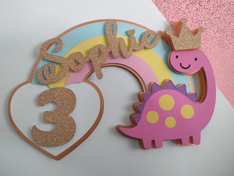 May include: A layered cake topper featuring a pastel rainbow, a white heart with a glittery gold number 3, and a pink dinosaur wearing a glittery gold crown. The name Sophie is written in glittery gold script across the rainbow arch.