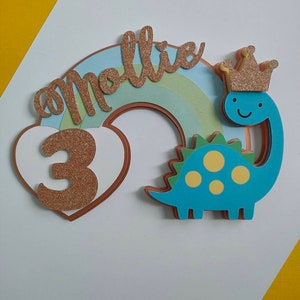 May include: A layered cake topper featuring a blue dinosaur with yellow spots wearing a gold glitter crown, a pastel rainbow, a white heart, and the name mollie in gold glitter script with the number 3 in gold glitter on the heart.