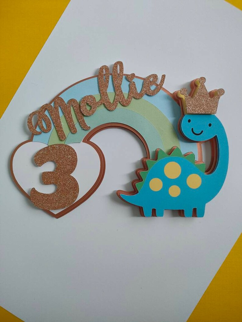 May include: A layered cake topper featuring a blue dinosaur with yellow spots wearing a gold glitter crown, a pastel rainbow, a white heart, and the name mollie in gold glitter script with the number 3 in gold glitter on the heart.