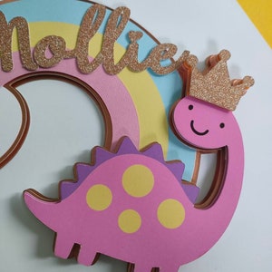 May include: A layered cake topper featuring a pink dinosaur with yellow spots and a gold glitter crown. The dinosaur is positioned in front of a pastel rainbow arch. The name Mollie is written in gold glitter script lettering across the rainbow.