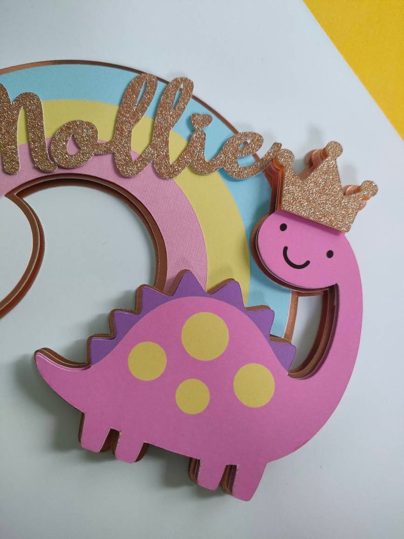May include: A layered cake topper featuring a pink dinosaur with yellow spots and a gold glitter crown. The dinosaur is positioned in front of a pastel rainbow arch. The name Mollie is written in gold glitter script lettering across the rainbow.