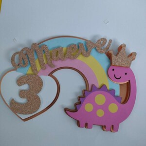 May include: A layered cake topper featuring a pastel rainbow, a pink dinosaur with yellow spots wearing a gold glitter crown, and a white heart. The name Maeve and the number 3 are written in gold glitter script and block lettering respectively.