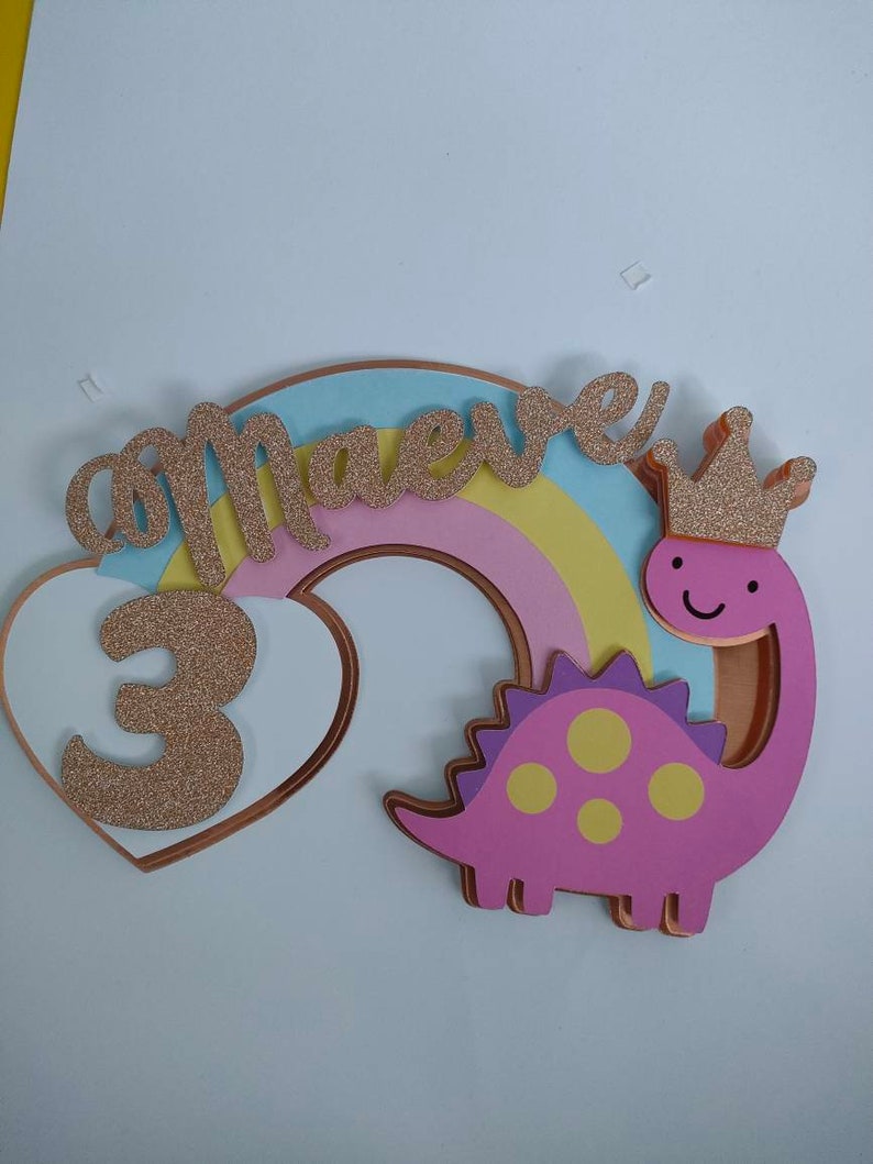 May include: A layered cake topper featuring a pastel rainbow, a pink dinosaur with yellow spots wearing a gold glitter crown, and a white heart. The name Maeve and the number 3 are written in gold glitter script and block lettering respectively.