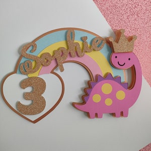 May include: A decorative cake topper featuring a pastel rainbow, a pink dinosaur wearing a gold glitter crown, and a white heart containing the number 3 in gold glitter. The name Sophie is written in gold glitter script across the rainbow arch.