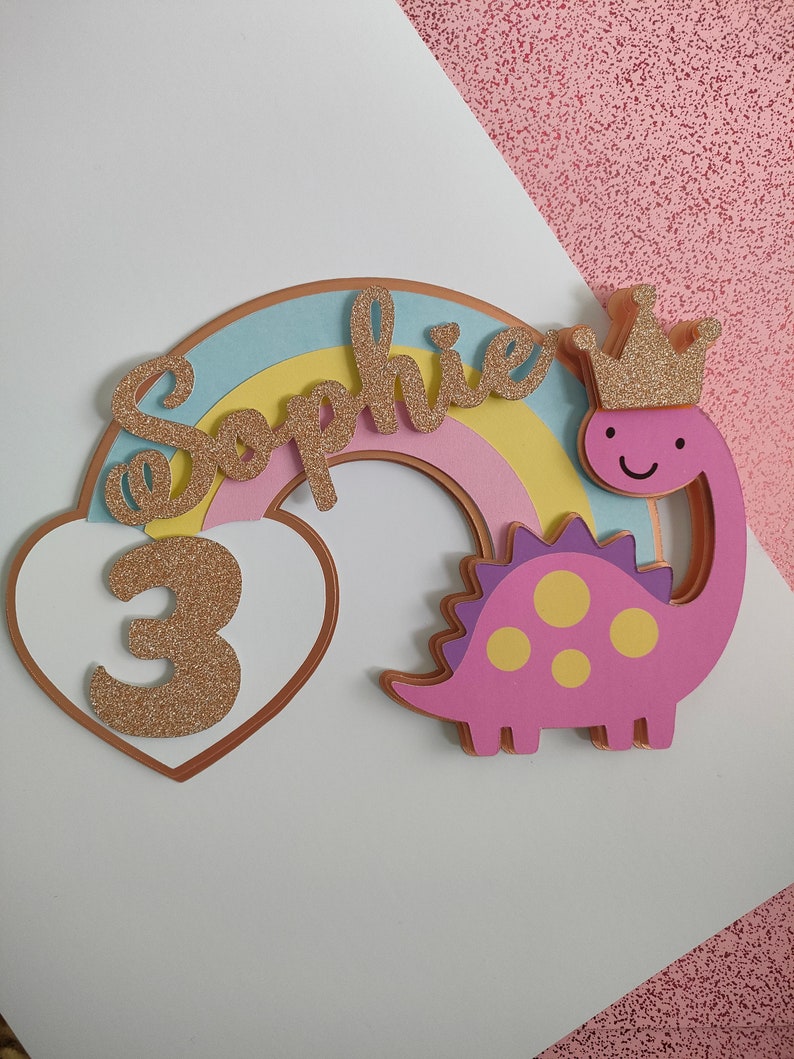 May include: A decorative cake topper featuring a pastel rainbow, a pink dinosaur wearing a gold glitter crown, and a white heart containing the number 3 in gold glitter. The name Sophie is written in gold glitter script across the rainbow arch.