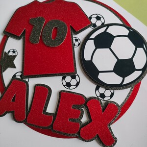 May include: Red and black soccer-themed cake topper with the name "ALEX" and the number "10" on a red jersey. The topper features a black and white soccer ball.