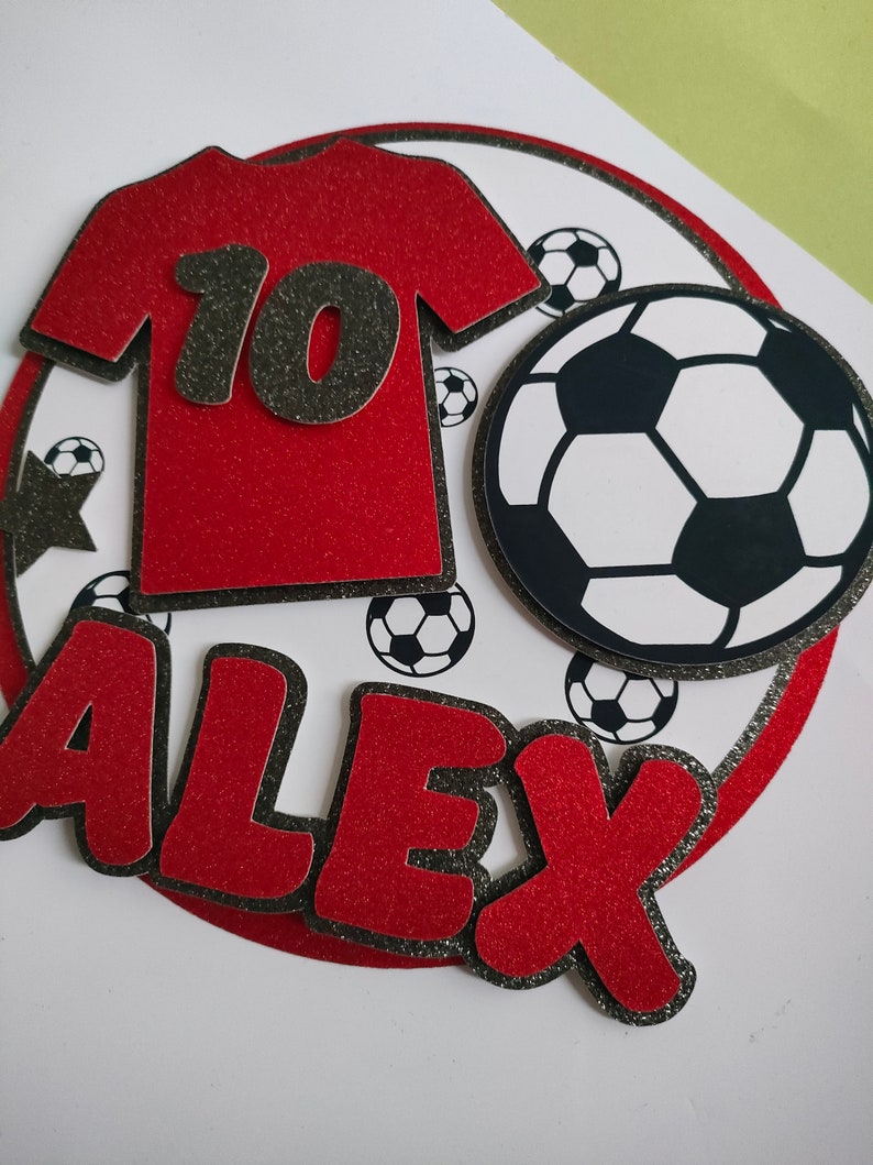 May include: Red and black soccer-themed cake topper with the name "ALEX" and the number "10" on a red jersey. The topper features a black and white soccer ball.
