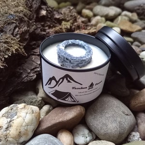 Könnte beinhalten: Eine schwarze Metall-Kerzendose mit einem weißen Etikett mit einem Berg- und Zeltmotiv. Die Kerze hat ein graues ringförmiges Element oben. Die Dose ist geöffnet und zeigt die Kerze im Inneren. Der Hintergrund umfasst Steine und Holz.