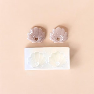 Peut inclure: Deux boucles d'oreilles en résine en forme de coquillage avec une découpe en forme de cœur, et un moule en silicone blanc avec deux cavités en forme de coquillage. Les boucles d'oreilles sont rose clair avec des paillettes, et le moule est blanc.