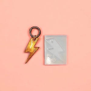 Peut inclure: Un charm en forme d'éclair doré pailleté avec un fermoir doré, à côté d'un moule en silicone transparent de la même forme. Le charm est un accessoire fait à la main, probablement pour des bijoux ou des porte-clés. Le fond est rose clair uni.