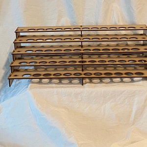 May include: A multi-tiered, unfinished wooden organizer with numerous circular holes designed to hold small bottles. The organizer has a stepped design, with each level slightly recessed. The wood has a natural, light brown color.