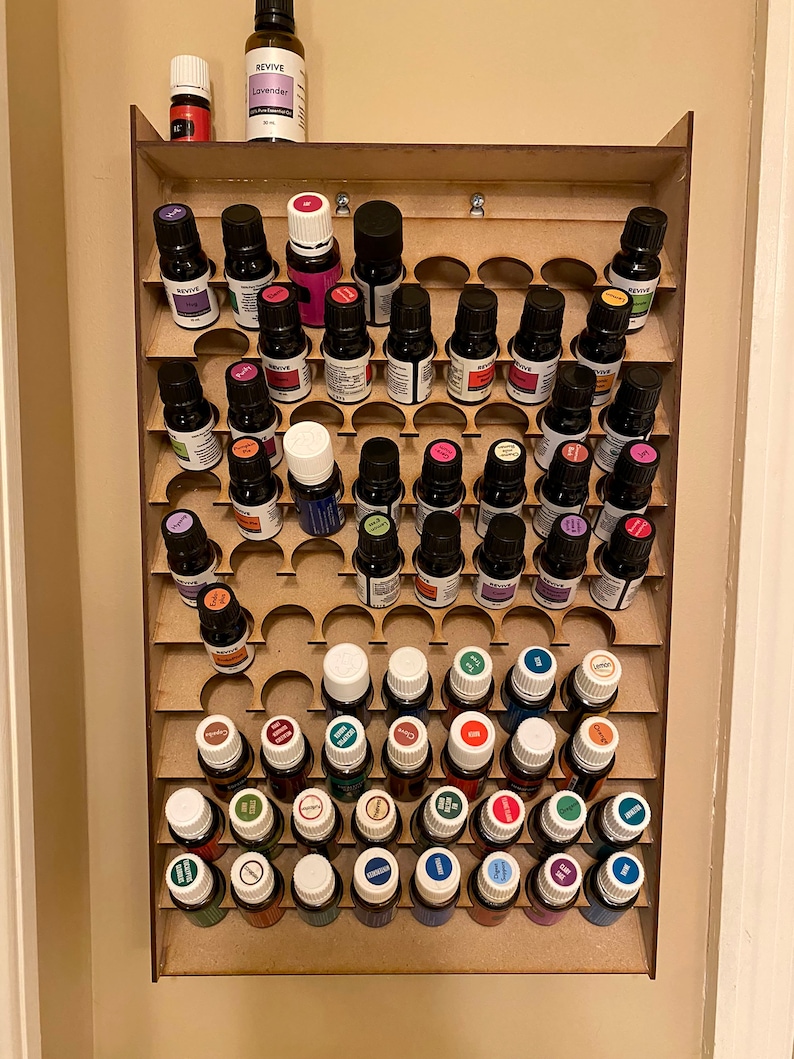 May include: A wooden essential oil organiser with multiple slots, holding numerous small glass bottles with colourful labels. The bottles are arranged in rows, showcasing a variety of essential oil products. The organiser is mounted on a wall.