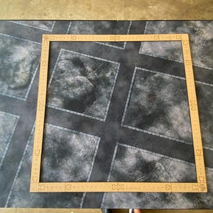 May include: A square, tan-colored wooden frame with etched markings rests on a gray and black patterned surface. The surface has a road and cobblestone design. The frame is likely for a tabletop game.