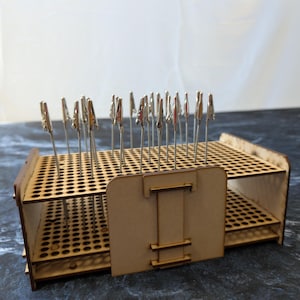 May include: A wooden paint handle holder with multiple holes for holding paint handles. The holder has two levels and a central support. Metal clips are inserted into the holes, ready to hold paint handles. The holder is made of light brown wood.