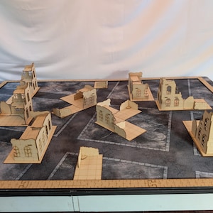 May include: A tabletop game scene featuring laser-cut wooden building ruins. The structures, in various states of disrepair, are arranged on a gray, grid-patterned surface, suggesting a post-apocalyptic or war-torn setting. The buildings have window cutouts.
