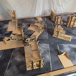 May include: A tabletop gaming terrain set featuring laser-cut wooden structures. The set includes various building ruins and modular terrain pieces, arranged on a gray and black surface. The pieces are light brown with intricate cut-out designs, suitable for miniature wargames and tabletop RPGs.