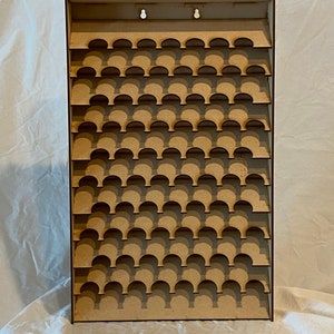 May include: A light brown wooden display shelf featuring numerous semi-circular cutouts for holding small objects. The rectangular shelf has a flat top with two holes for hanging. The design is simple and functional, suitable for displaying collectibles or miniatures.