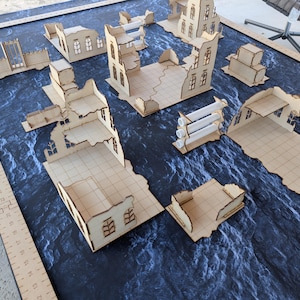May include: A collection of laser-cut light wood miniature building ruins for tabletop gaming, arranged on a dark blue textured mat. A wooden ruler along the left edge shows measurements in inches, with structures featuring grid floors and walls.